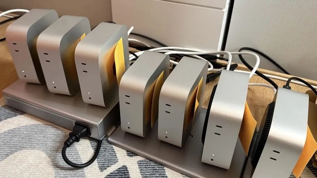 七台银色Mac Mini电脑并排竖立在两台笔记本电脑上，每台电脑上都贴着黄色便签，电源线和各种线缆缠绕在后面的木桌上。